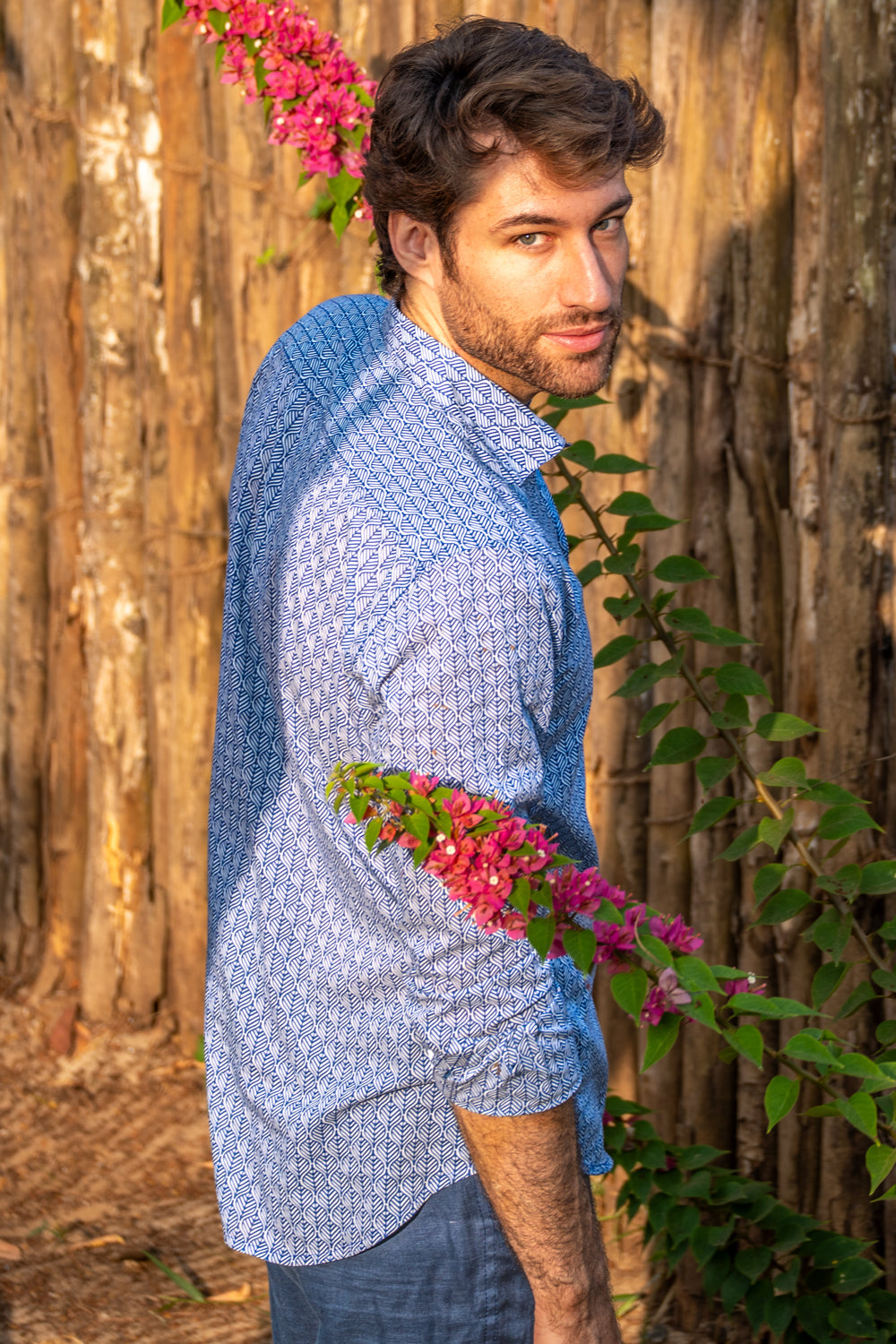 Man in a blue patterned shirt standing against a wooden fence with pink flowers.