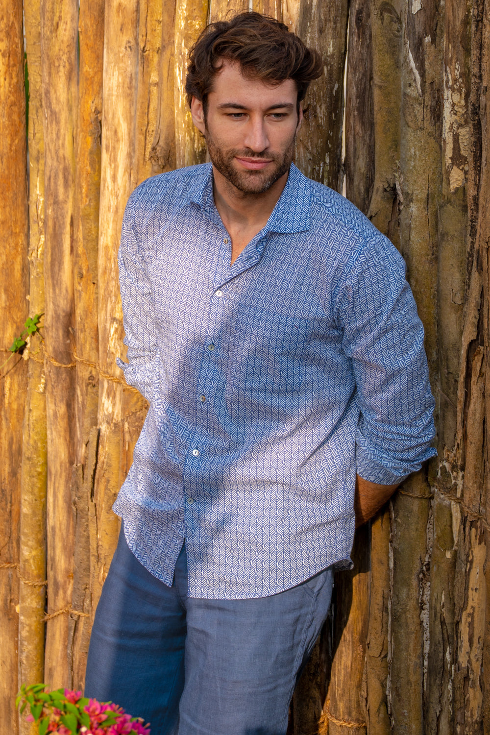 Man wearing a blue patterned shirt leaning against a wooden wall.